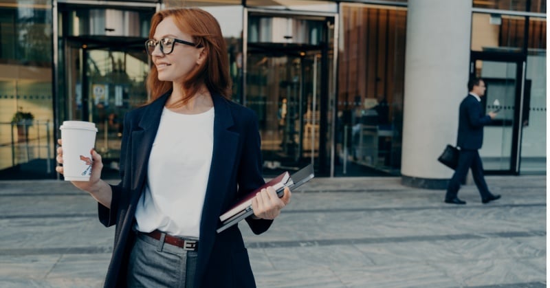 Professional woman outside an office building holding a coffee cup and folders while looking to the side, with another businessperson walking in the background.