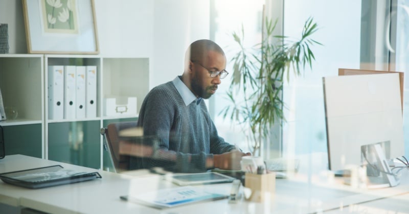 Man working at a desk in a bright modern office, viewed through glass, with a computer monitor, shelves of binders, and plants in the background.