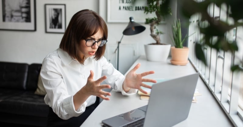 Frustrated woman in glasses gesturing at an open laptop while seated at a bright home office desk with plants and framed wall art in the background.