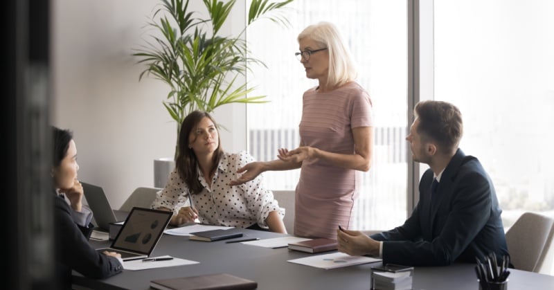 Four professionals in a conference room meeting, with one woman standing and speaking while others listen around a table with laptops and documents.