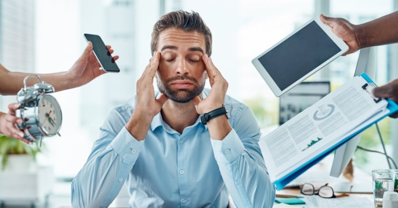 Overwhelmed office worker sitting at a desk with eyes closed and fingers on his temples while multiple hands around him hold a phone, alarm clock, tablet, and documents, symbolizing interruptions and workplace stress.
