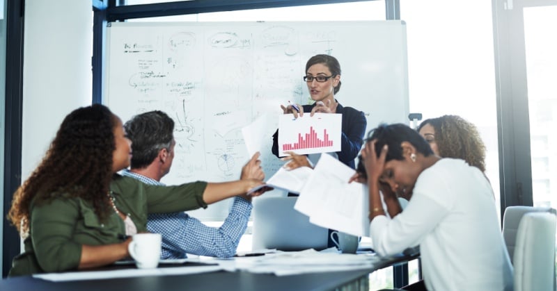 Team meeting with a manager presenting a chart at a whiteboard while colleagues appear confused and overwhelmed by papers, illustrating communication breakdown in the workplace.