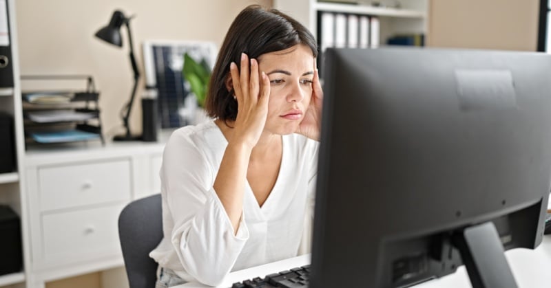 Woman sitting at a desk in front of a computer with her hands on her temples, looking stressed while working in a home office.