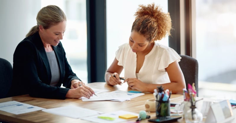 Two professionals seated at a desk reviewing paperwork together in a bright office.