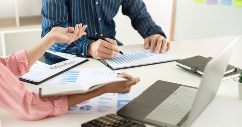 A close-up of two people reviewing financial charts and documents at a desk with a laptop, tablet, calculator, and notebook during a business meeting.