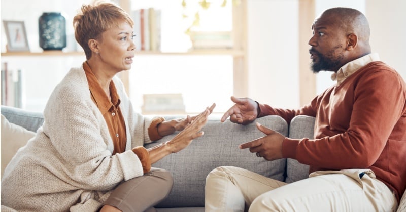 Couple arguing on a couch at home, gesturing with frustration during a tense conversation.