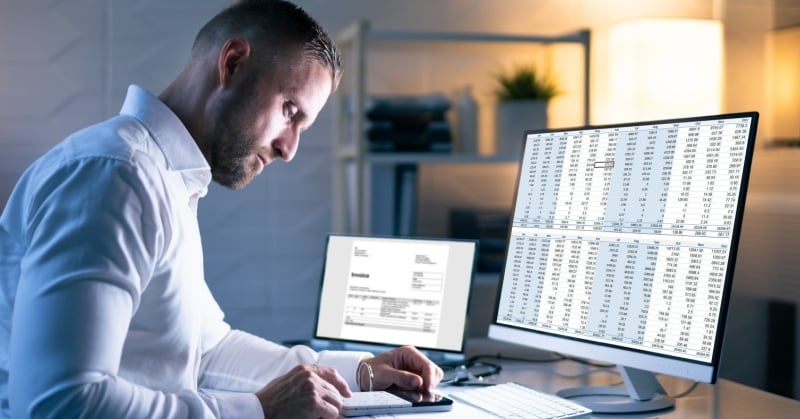 Accounting professional reviewing financial data on a desktop monitor while calculating figures at a desk, with an invoice displayed on a second screen.