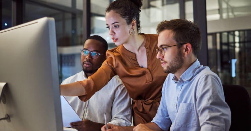Three professionals collaborate at a computer in a modern office, reviewing information together on a large monitor.