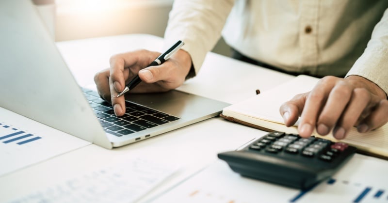 Person using a laptop and calculator while writing in a notebook at a desk with financial charts.