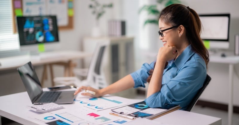 A woman reviews financial charts and reports at her desk while participating in a video meeting on her laptop.
