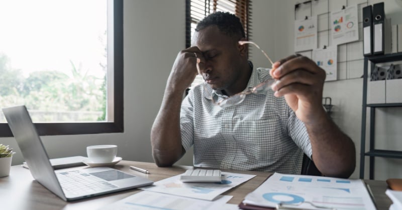 A man sits at a desk in an office, looking stressed while holding his glasses, with a laptop, papers, and charts spread out in front of him.