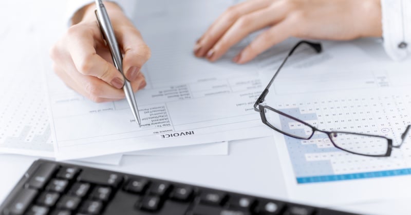 Close-up of a person’s hand holding a pen and writing on an invoice document, with eyeglasses resting on paperwork and a computer keyboard in the foreground.