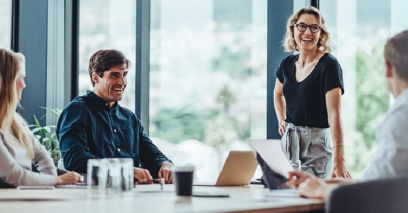 A diverse group of professionals engaged in a cheerful and collaborative meeting around a conference table, with a woman standing and smiling while others are seated and interacting in a modern office with large windows.