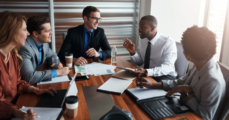 Diverse group of five business professionals meeting around a conference table with laptops, documents, and coffee cups, engaged in discussion in a modern office.