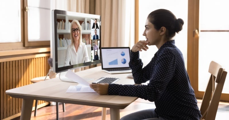 Woman seated at a home office desk during a video meeting, looking at a large monitor with several colleagues on screen while holding papers, with charts displayed on a laptop beside her.