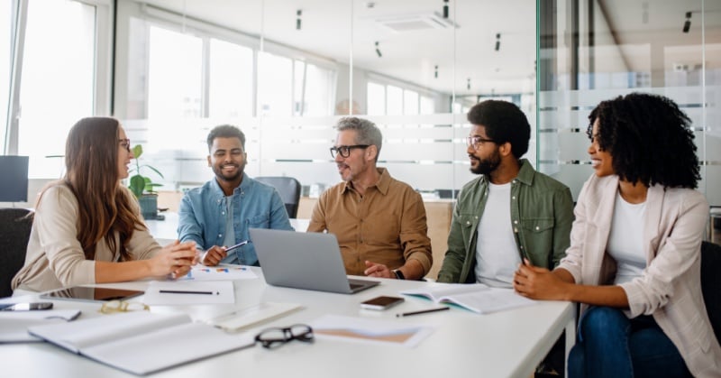 Five colleagues meeting around a conference table in a bright modern office, discussing documents and a laptop.