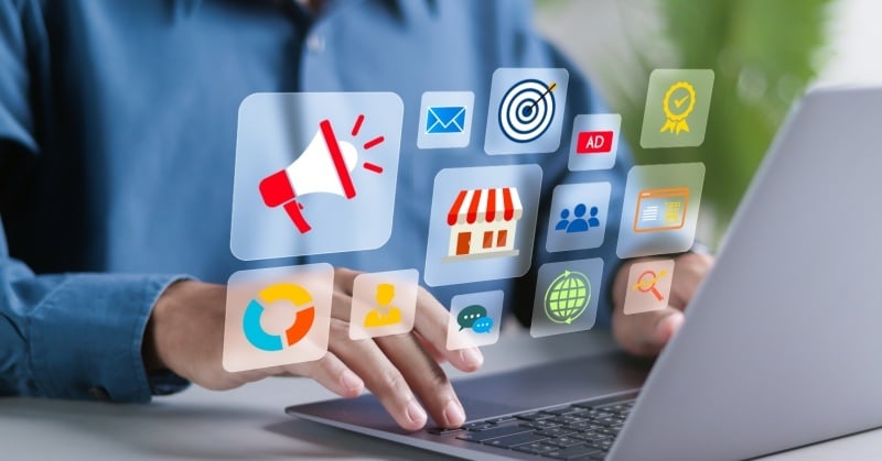 Person using a laptop with floating digital marketing icons, including email, advertising, analytics, search, and storefront symbols.