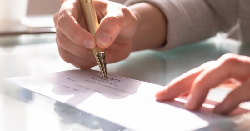 A close-up of a person's hand holding a gold pen and writing a check on a reflective glass surface, suggesting a financial or payment-related activity.