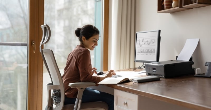 Young professional woman working remotely from a home office, smiling and engaged with financial charts on a desktop monitor, seated at a modern ergonomic chair by a window.