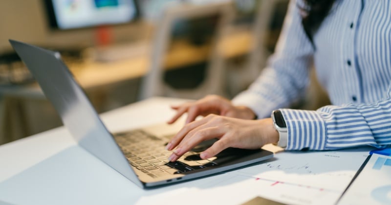 Woman typing on a laptop at a desk with financial charts and reports spread out nearby.
