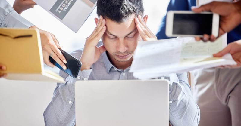 "A stressed professional man sits at a desk with his hands on his temples, surrounded by multiple outstretched hands offering documents, folders, a smartphone, and a tablet, conveying a sense of overwhelming demands and workplace stress.