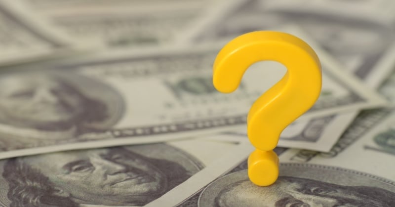 A yellow question mark stands upright on a background of scattered U.S. one hundred dollar bills, symbolizing financial uncertainty.