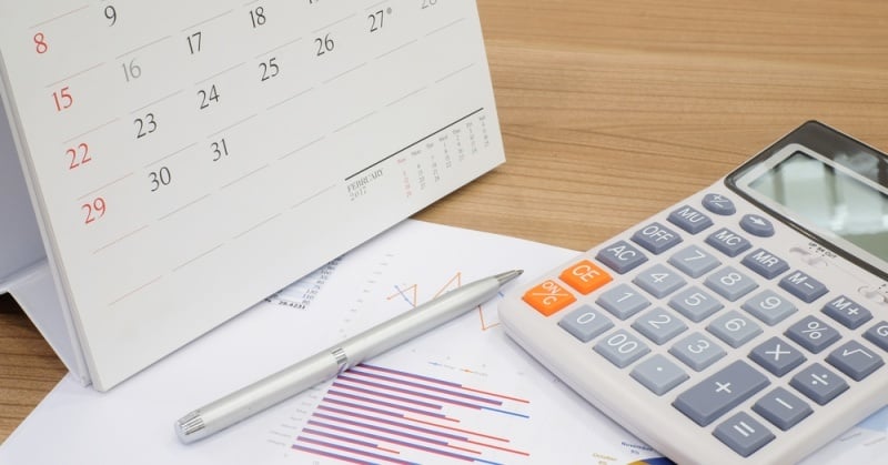 Desk scene with a standing paper calendar open to February, financial documents with bar and line charts, a silver pen, and a large calculator.