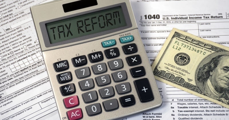 A close-up image of a calculator with the words "TAX REFORM" displayed on its screen, placed on top of U.S. tax forms, including IRS Form 1040, with a one hundred dollar bill partially visible next to it.
