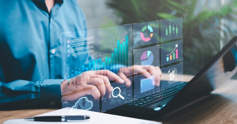A businessperson types on a laptop at a wooden desk with floating holographic graphs and charts representing financial analytics, set in a modern office environment.