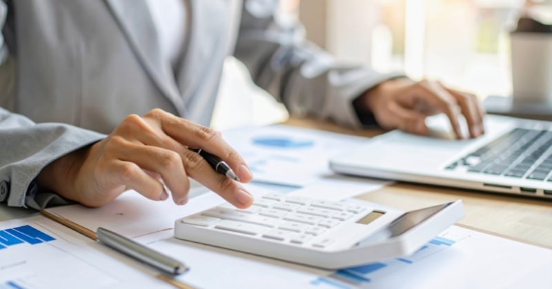 Accountant using a calculator and laptop while reviewing financial charts and reports.