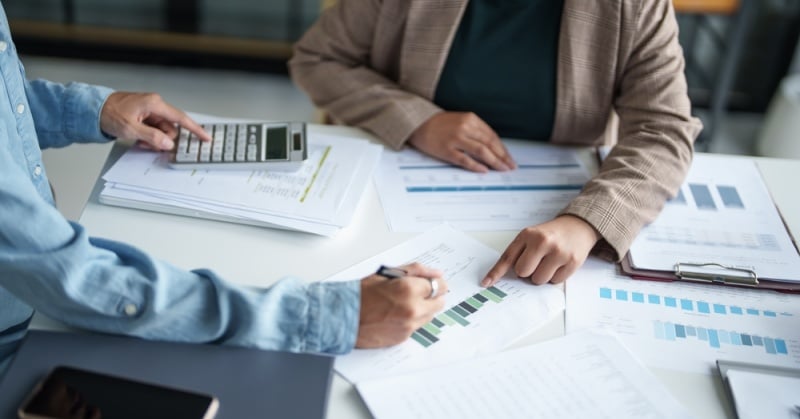 Two professionals reviewing financial documents and charts at a desk, using a calculator and discussing tax planning strategies for business entities. 