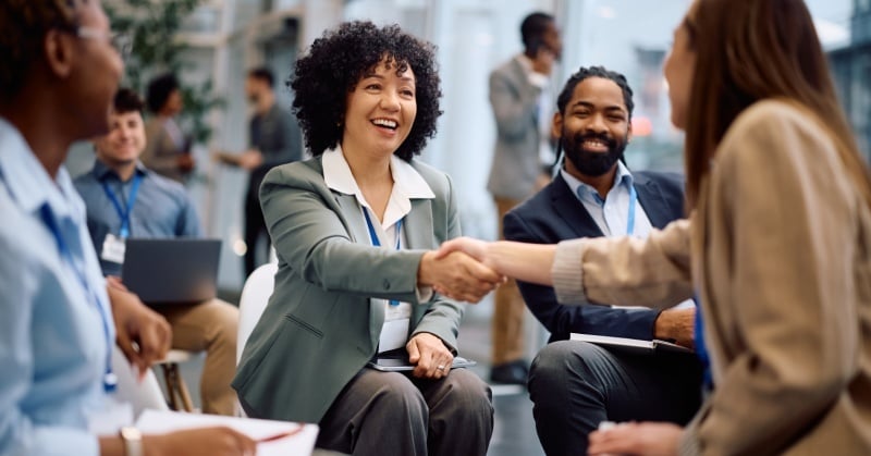 A diverse group of professionals attend a networking event, with two attendees smiling and shaking hands during a seated conversation.