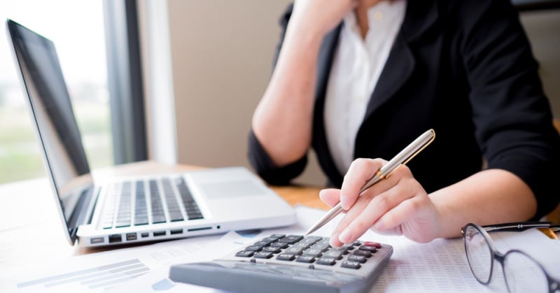 Professional bookkeeper at a desk using a calculator and referencing financial documents, with a laptop in the background.