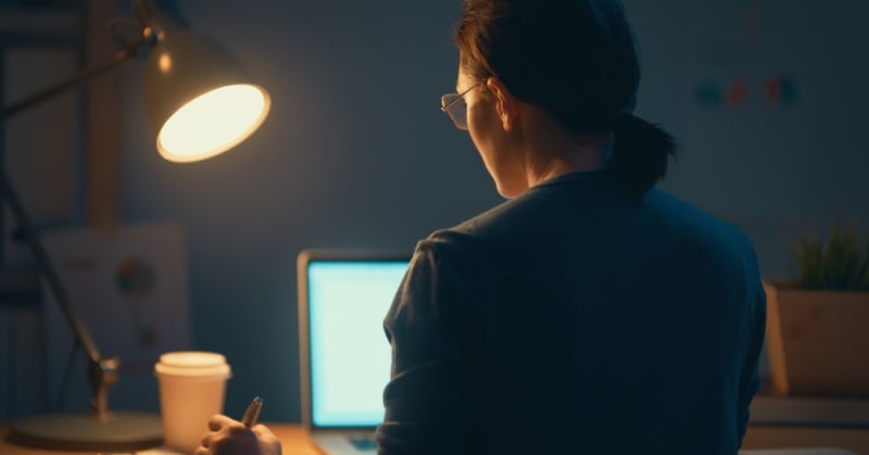 A person working alone at a desk late at night, illuminated by a desk lamp, with a coffee cup beside a laptop symbolizing isolation and late hours often experienced by accounting professionals.