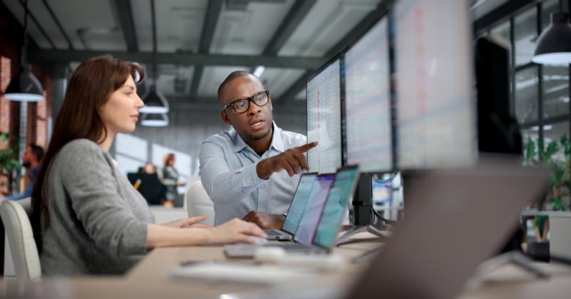 Two accounting professionals review data across multiple computer monitors in a modern office setting.