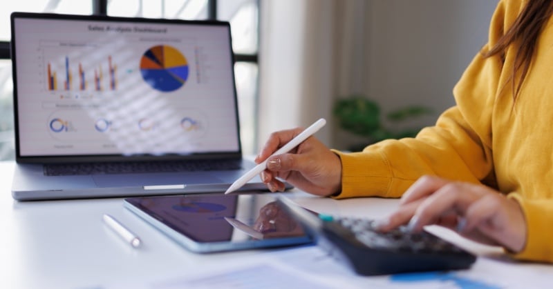 Person using a stylus and calculator while reviewing financial charts and graphs on a laptop and tablet.