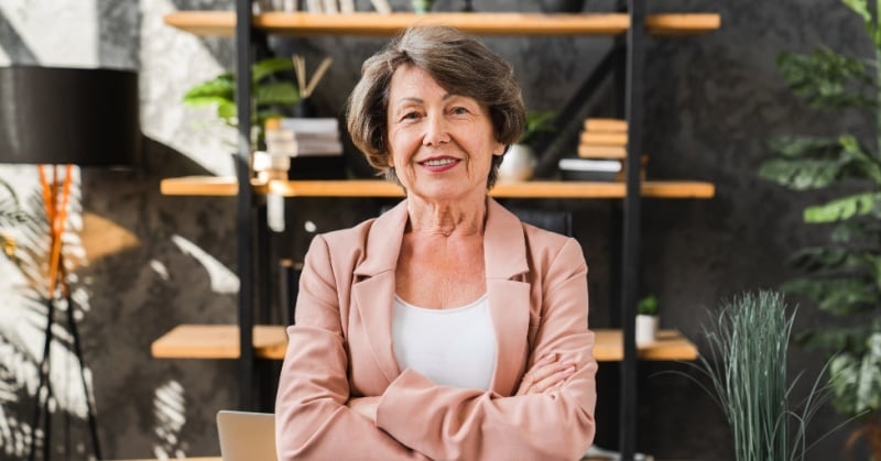 A confident older woman with short gray hair, wearing a light pink blazer over a white top, stands with arms crossed in a modern office setting with bookshelves, plants, and soft natural lighting in the background.