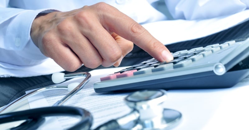 A healthcare professional uses a calculator on a desk with medical equipment, including a stethoscope, in the background, symbolizing financial management in healthcare.
