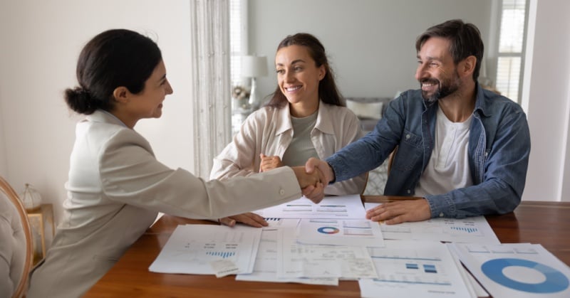 Professional advisor shaking hands with clients during a financial planning meeting, with charts and reports spread across the table.