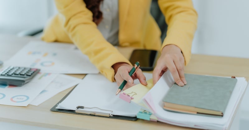 Person taking notes in a planner beside financial reports, a calculator, and a notebook.