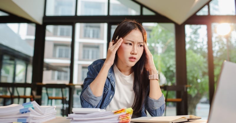 Woman sits at a desk with stacks of paperwork, holding her head with a stressed expression.