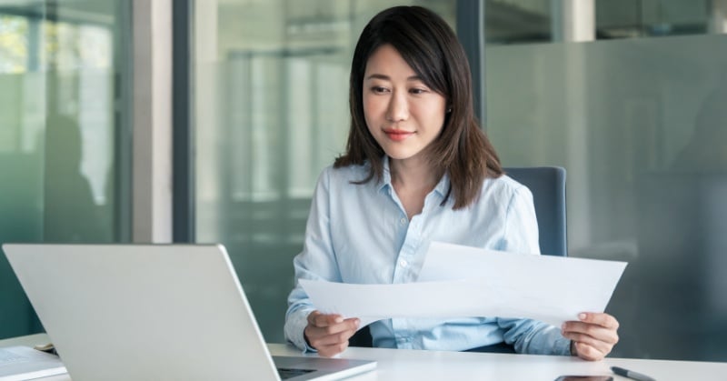 A professional woman sits at a desk in a modern office, holding printed documents and looking attentively at a laptop screen.