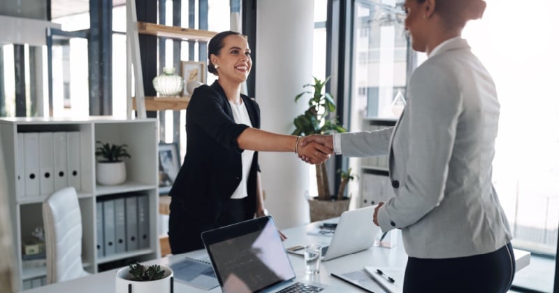 Two professional women in business attire shake hands in a bright, modern office setting.