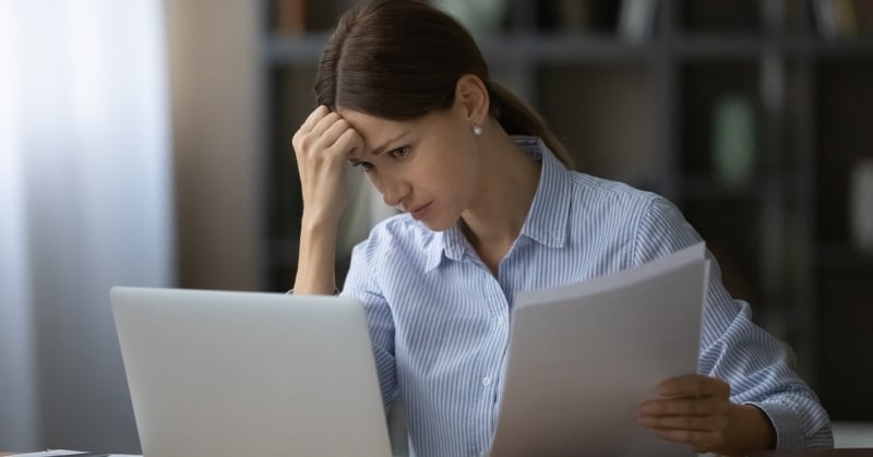 Woman reviewing paperwork beside a laptop with a worried expression.