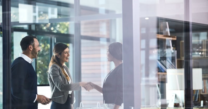 Three professionals are seen through a glass wall in a modern office setting, with two women shaking hands and a man smiling beside them, suggesting a business introduction or agreement.