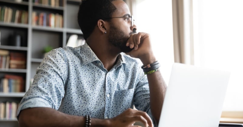 Man sitting at a desk with a laptop, gazing thoughtfully out a window with bookshelves in the background.