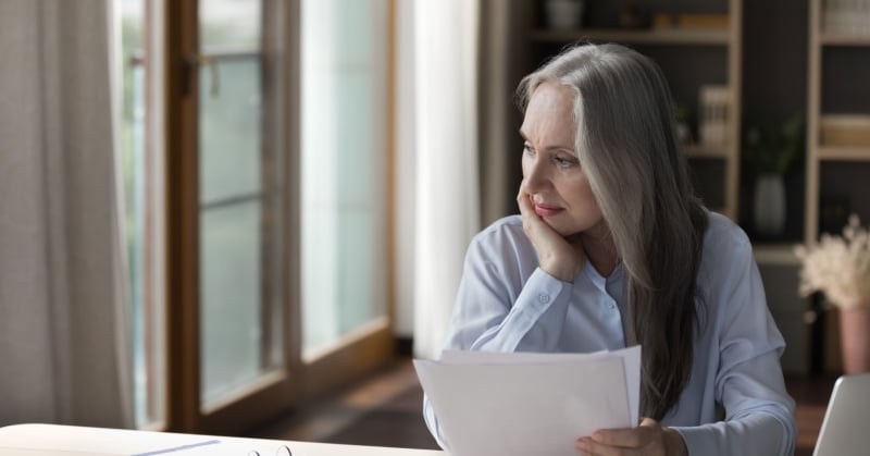 Older woman with long gray hair sitting at a desk holding documents, looking thoughtfully out a window in a home office setting.