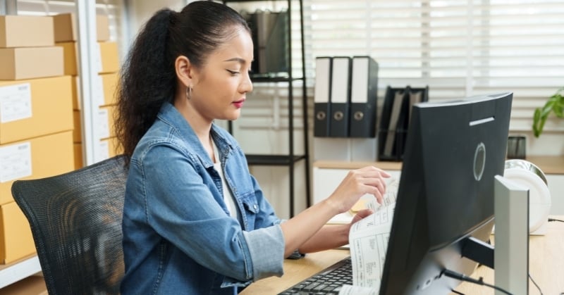 Woman reviewing inventory documents at a computer in a small e-commerce warehouse, with shipping boxes and office supplies in the background.