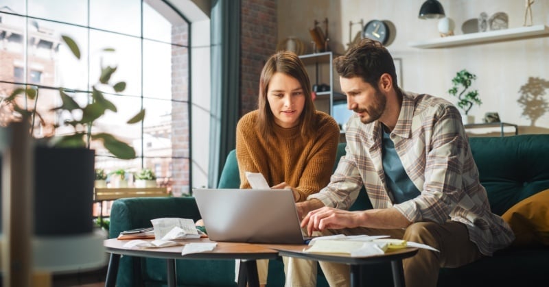 A couple sits together at a coffee table in a modern living room, reviewing receipts and using a laptop, suggesting financial planning or document organizatio
