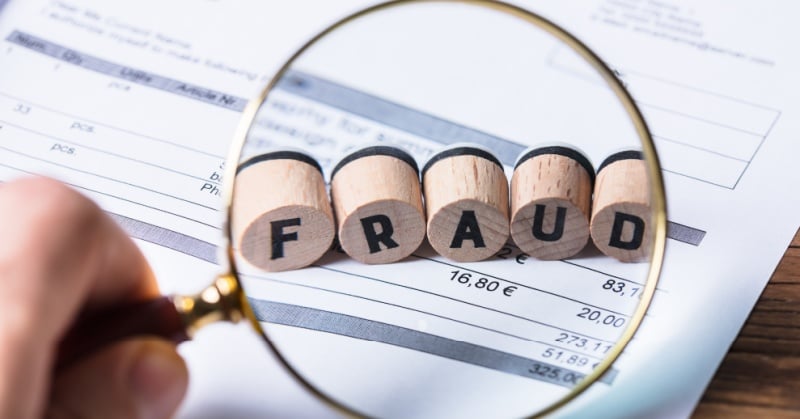 A magnifying glass hovers over wooden blocks spelling "FRAUD" on top of a financial document, symbolizing the scrutiny and detection of financial misconduct.